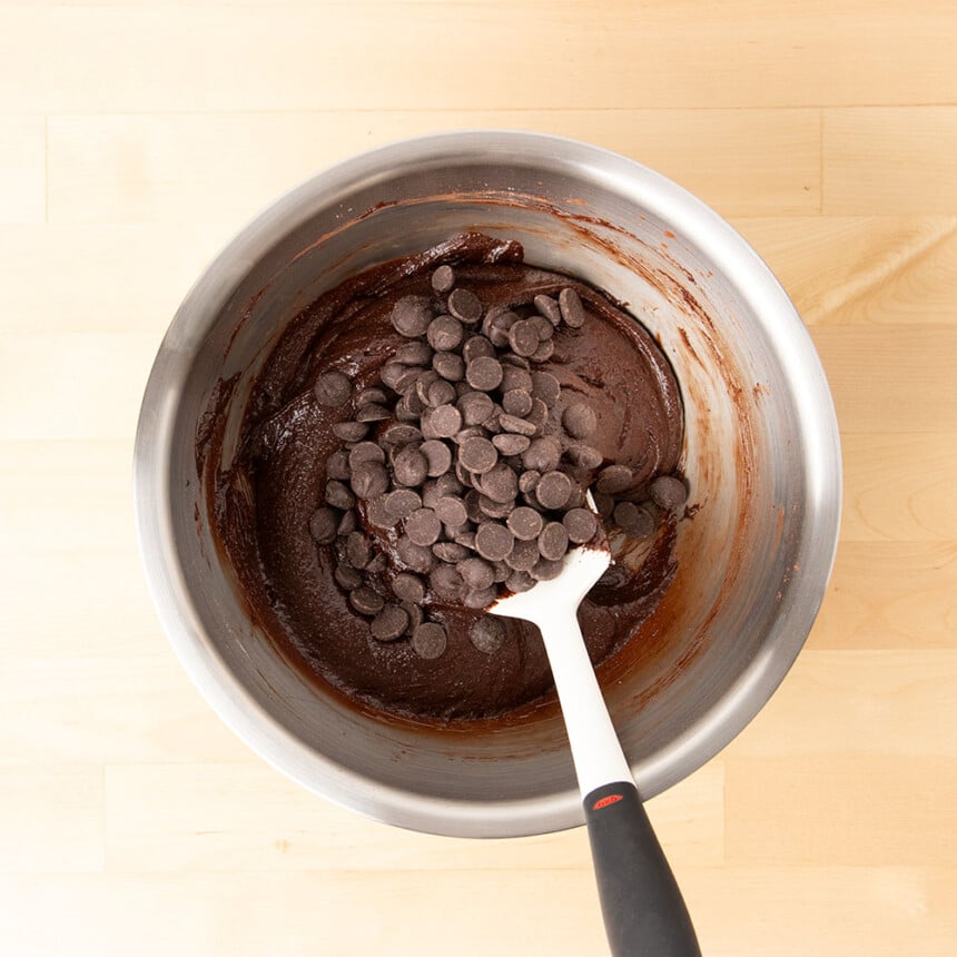 chocolate chips on top of dark brown brownie batter with white spatula in mixing bowl