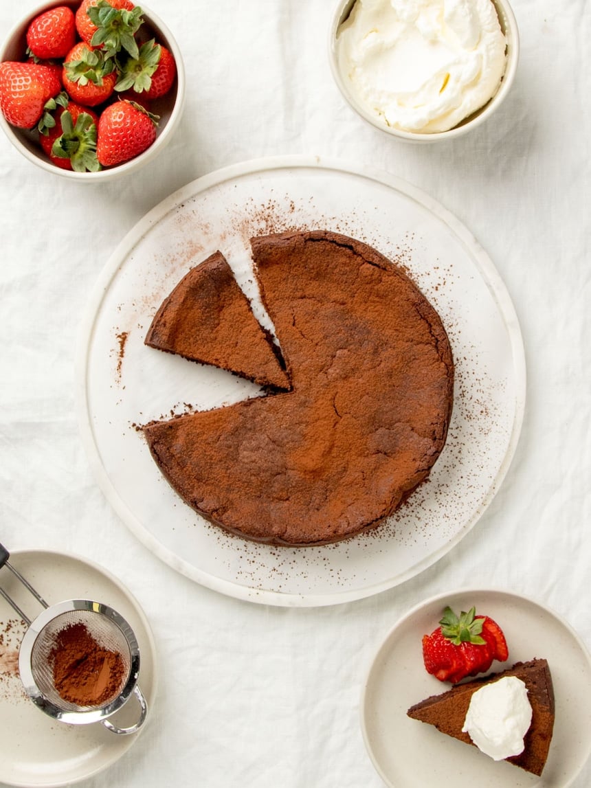 overhead image of brown chocolate cake with cocoa powder with one slice cut and one slice missing, slice on small plate with shipped cream, strawberries in bowl, whipped cream in bowl