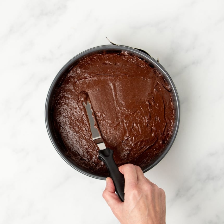 hand using small metal offset spatula with black handle to spread out raw flourless chocolate cake batter in same round springform pan