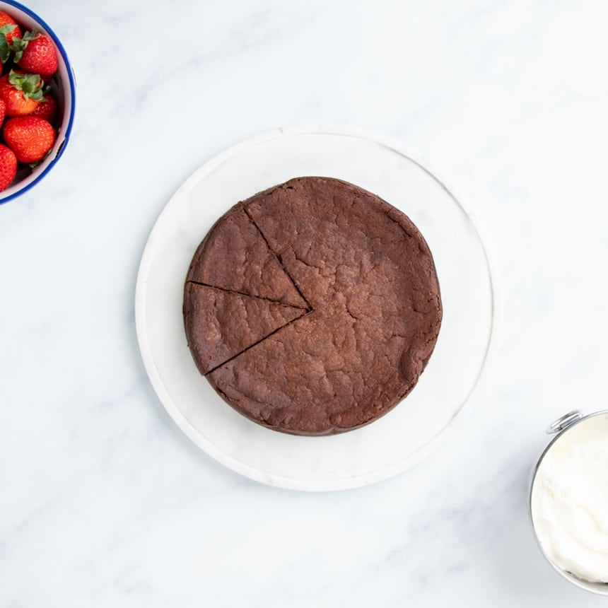 overhead image of brown flourless chocolate cake with 2 slices cut into cake; cake sitting on white flat platter with small white bowl of strawberries and small metal bowl of whipped cream