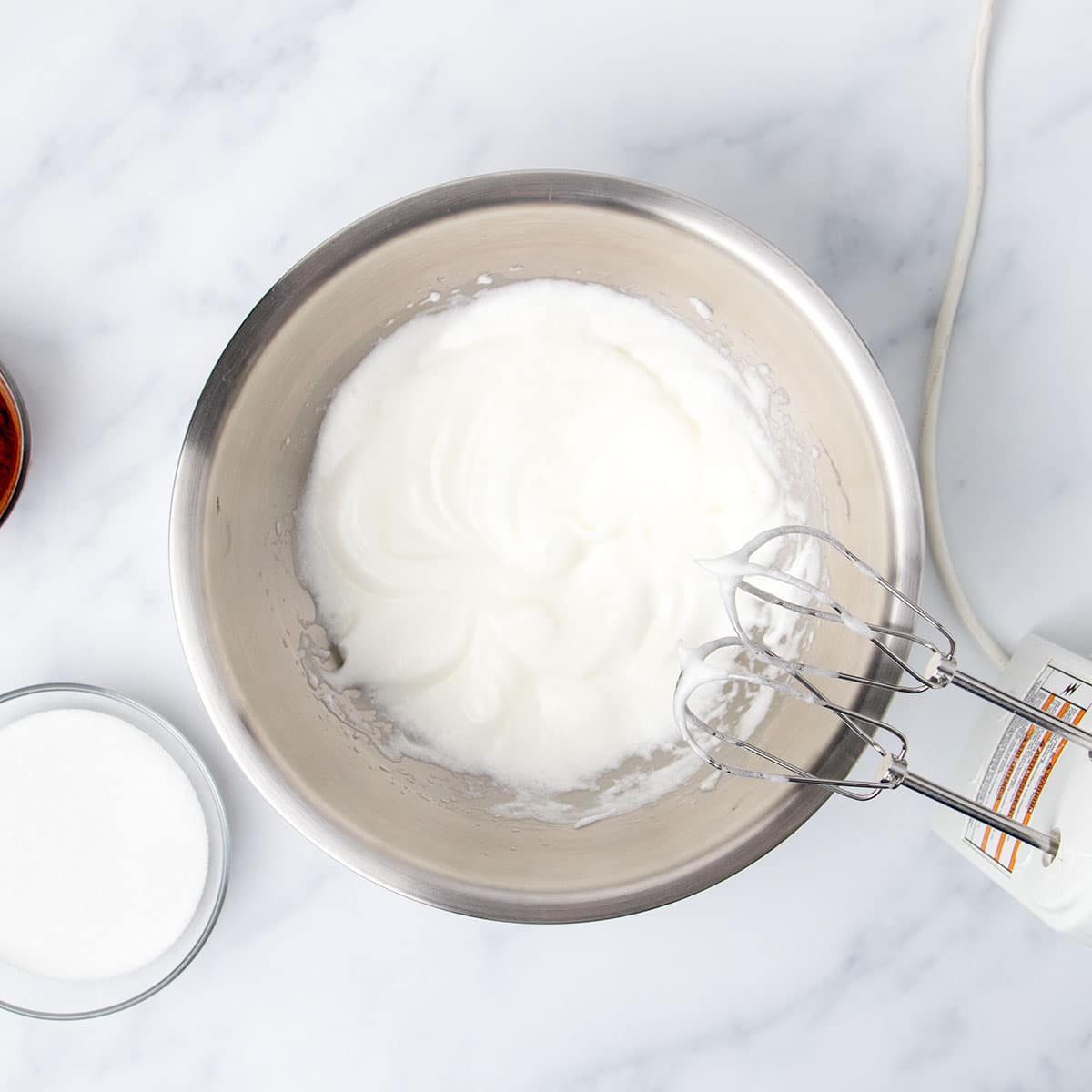 Whipped egg whites in large round metal mixing bowl with white handheld mixer.