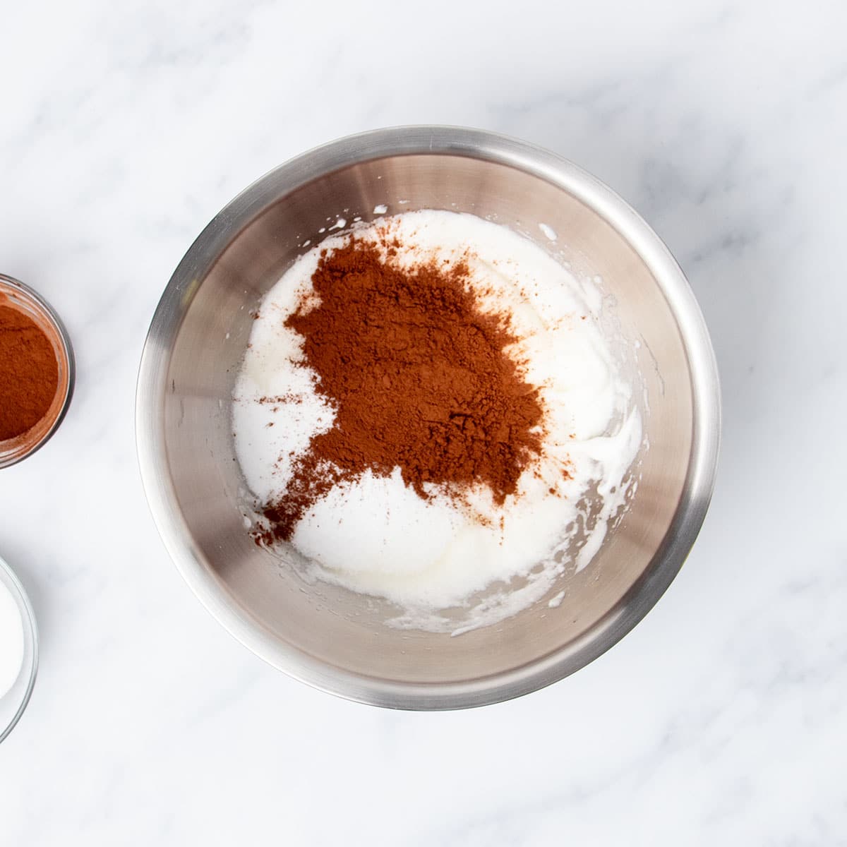 Cocoa powder and sugar sprinkled on top of whipped egg whites in same metal mixing bowl.