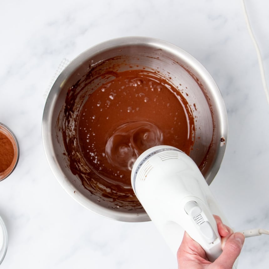 hand holding white handheld mixer beating egg whites with cocoa powder and sugar