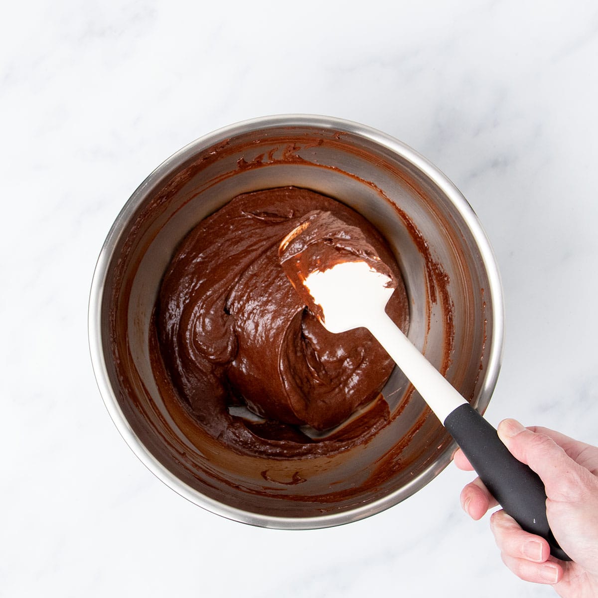 hand holding white spatula folding in chocolate meringue mixture into whisked ingredients in same bowl