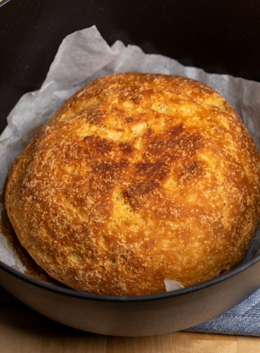 light brown crusted round gluten free artisan bread in round black dutch oven pot with white parchment paper underneath and pot on blue cloth