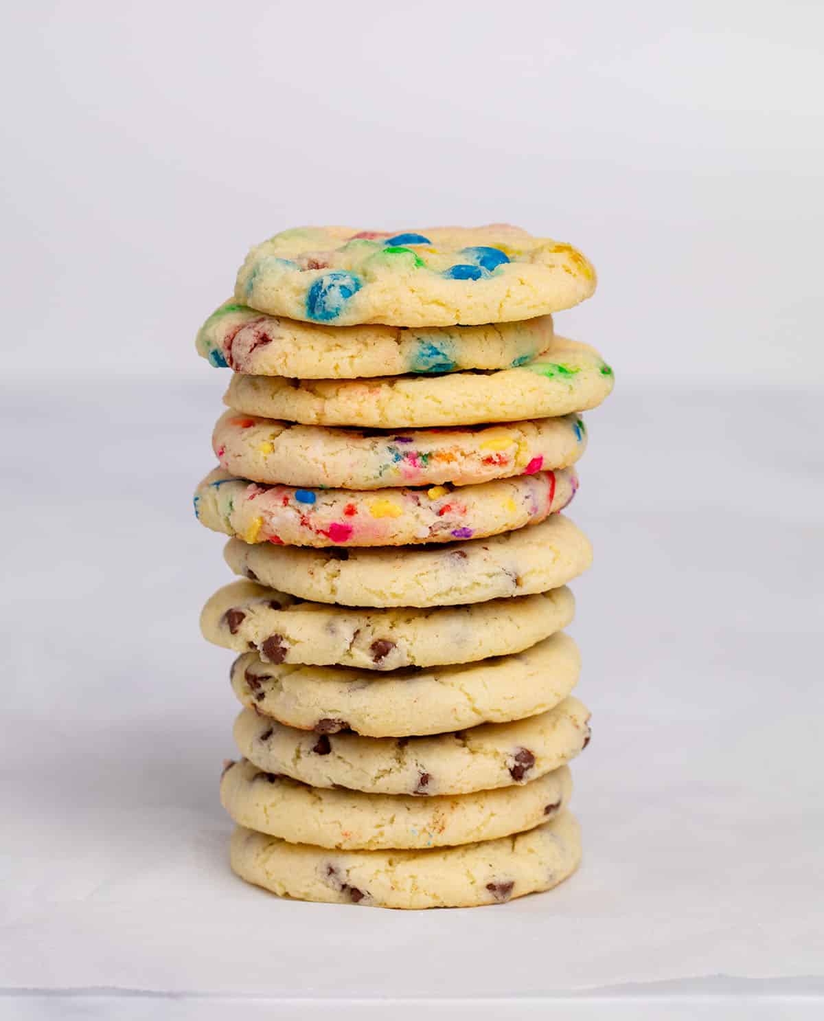 Tall stack of 11 gluten free cake mix cookies with different mix ins on white paper.