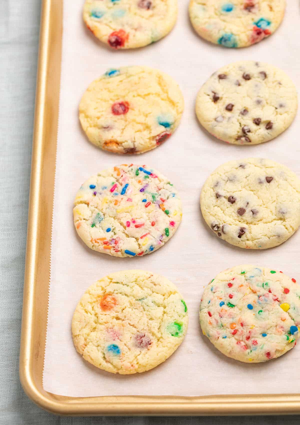 8 baked cookies with different mix ins baked on white paper-lined gold rimmed baking sheet on blue cloth.