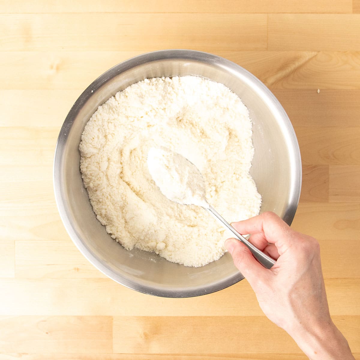 Hand holding metal mixing spoon with butter mixed into dry ingredients in same metal mixing bowl.