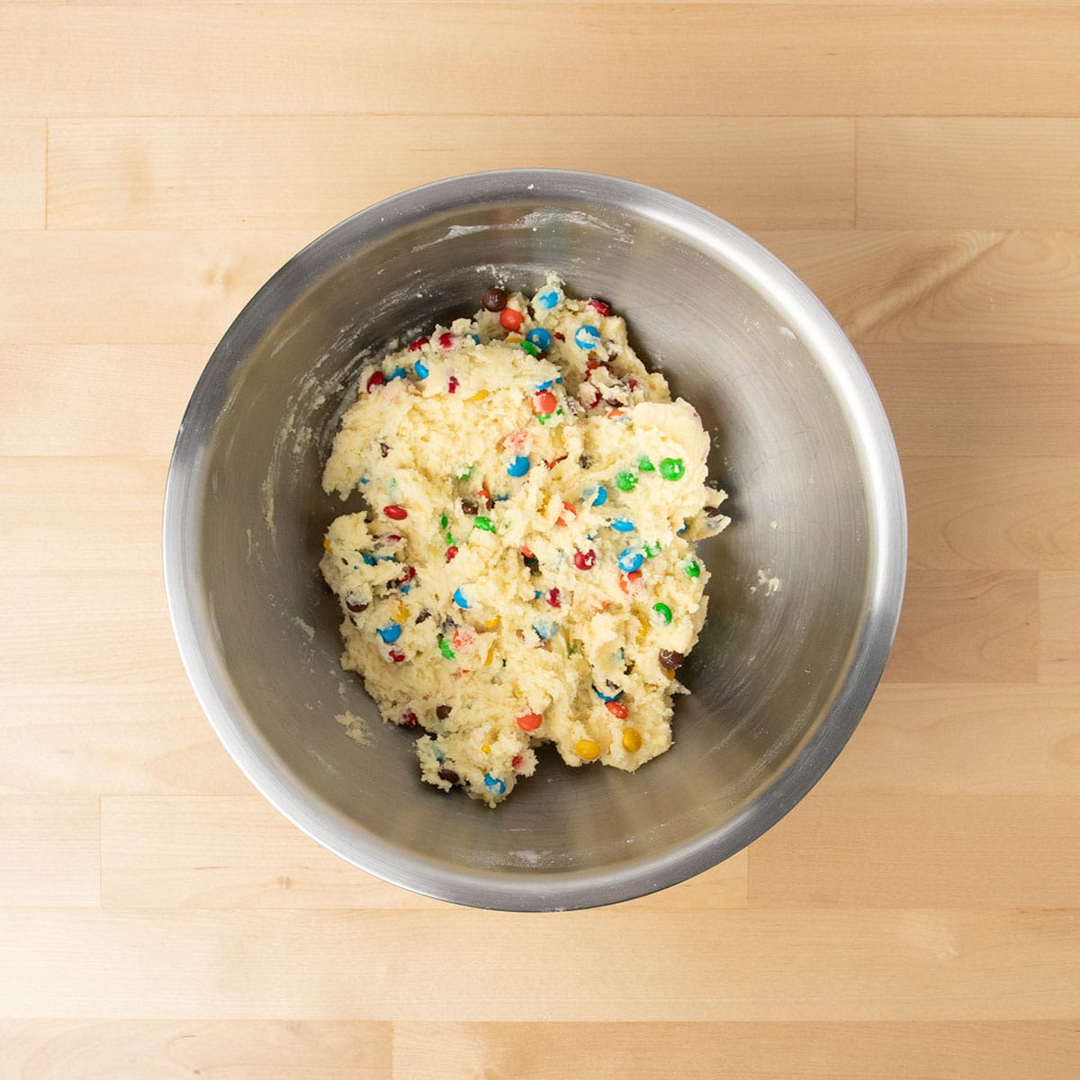 Colored candy pieces mixed into light yellow cookie dough in mixing bowl.