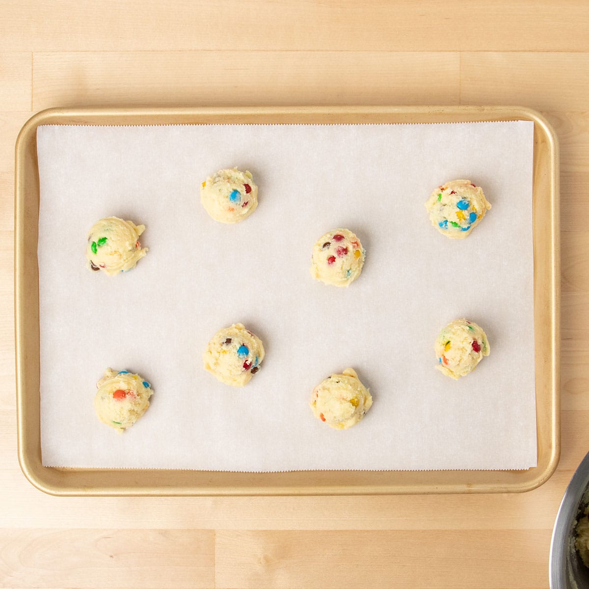 8 small scoops of light yellow cake mix cookie dough with colored candy pieces on white paper on baking tray.