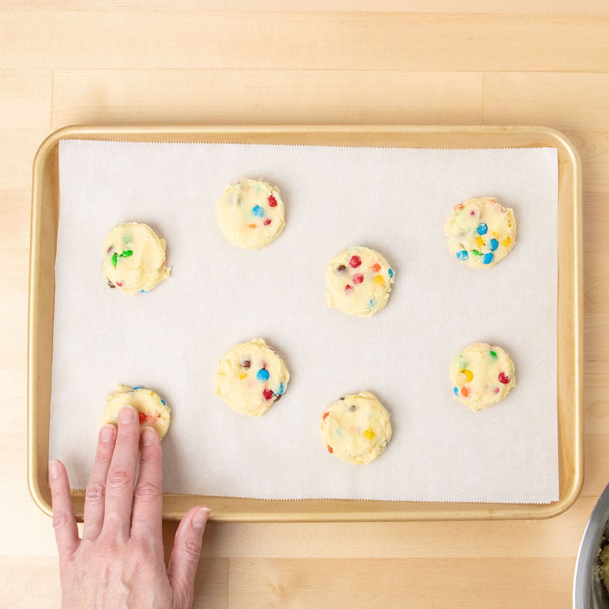 Cookie dough pieces with fingers pressing them into disks on white paper on baking tray.