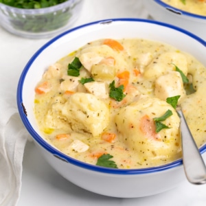 gluten free chicken and dumplings closeup image of 2 dumplings with soup, chicken, carrots and herbs in white bowl with flat leaf parsley scattered