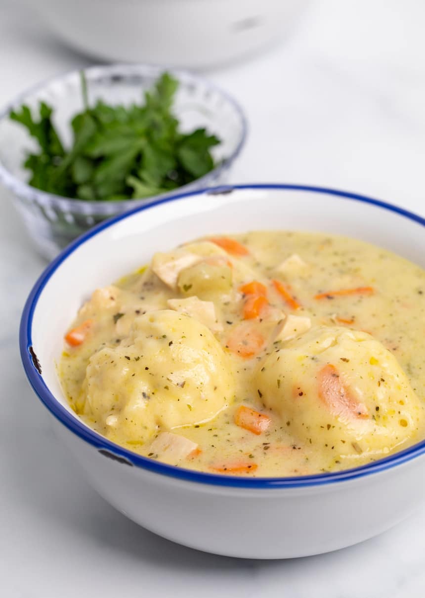 gluten free chicken and dumplings in white bowl with blue rim with green parsley in background