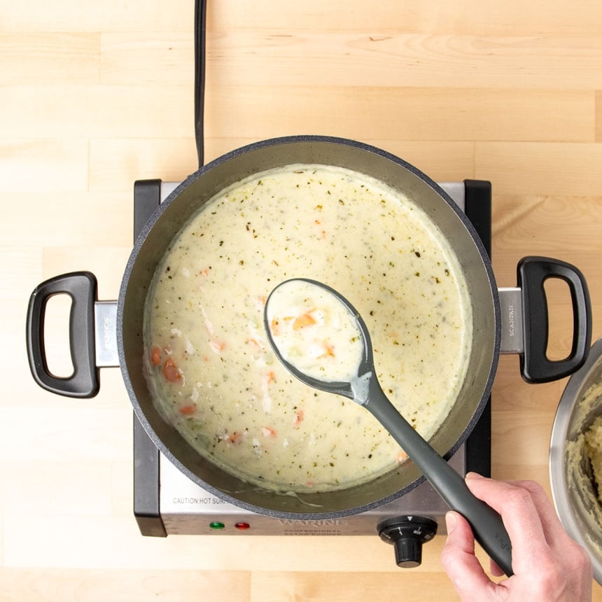 hand holding large black mixing spoon with spoonful of light tan cream soup with carrots and celery in pot