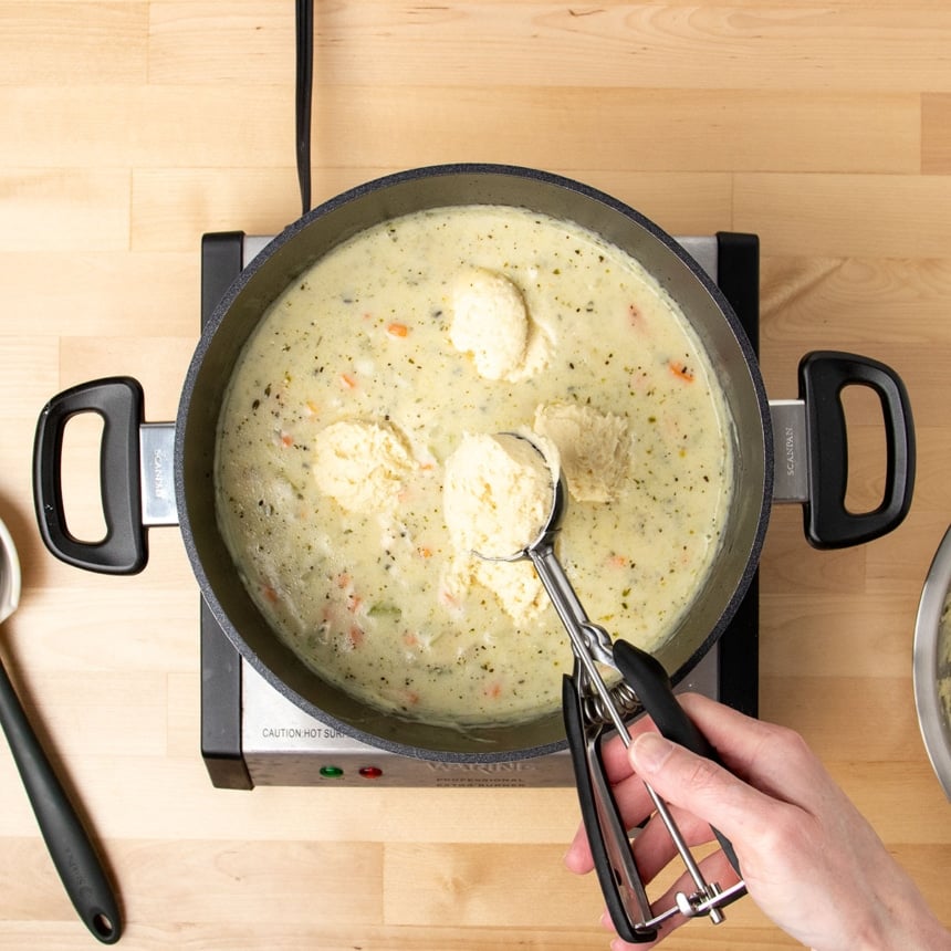 hand holding ice cream scoop with black handle releasing raw dumpling batter into pot of soup