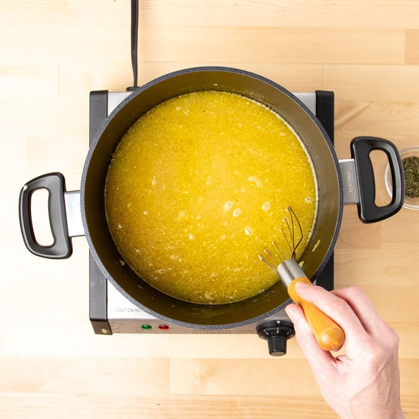 hand holding flat whisk with wooden handle in same pot with yellow chicken stock