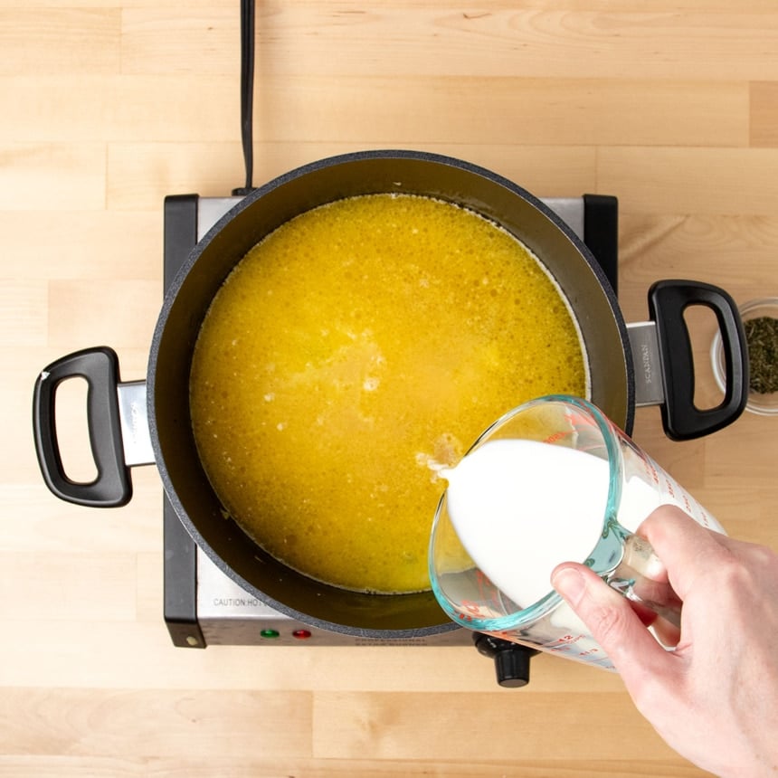 hand pouring white milk from glass measuring cup into black pot with chicken stock