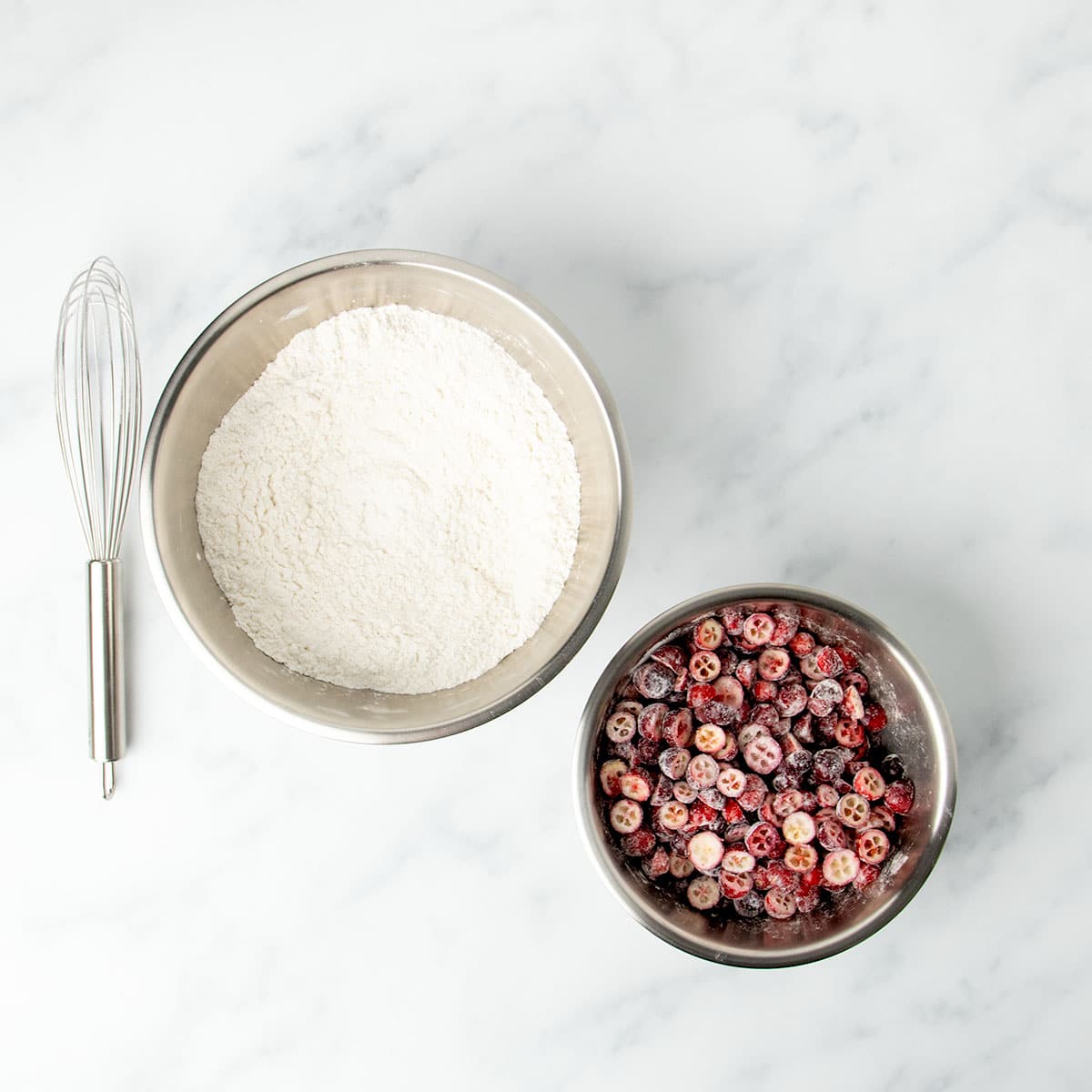 Large metal mixing bowl with dry ingredients, large wire whisk, and small metal mixing bowl with halved cranberries tossed white powder, all on marble surface