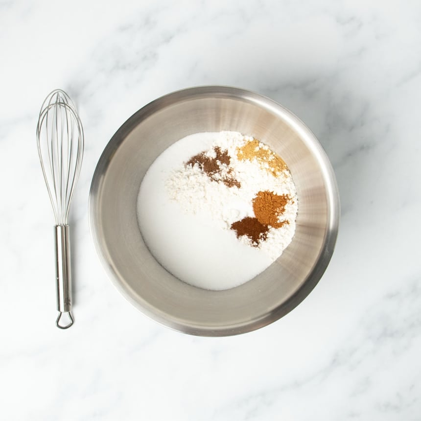 large round metal mixing bowl with gluten free flour blend, white sugar, and dark and light brown spices with large wire whisk next to bowl on marble surface