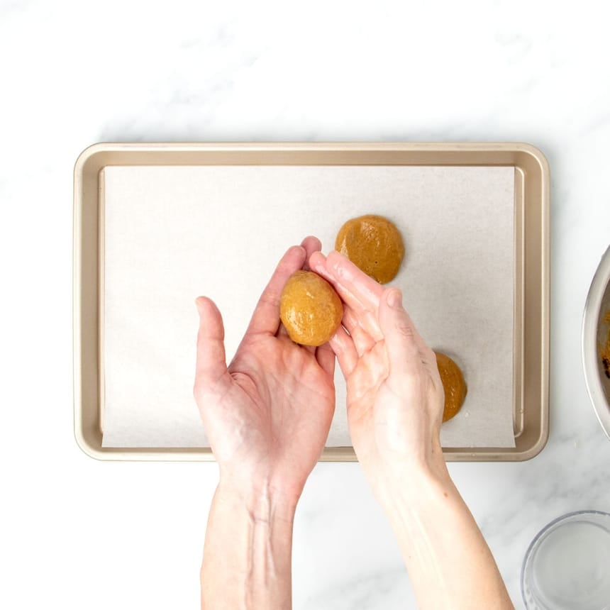 moistened fingers of two hands holding and shaping a soft ball of molasses cookie dough above a white paper-lined gold baking sheet