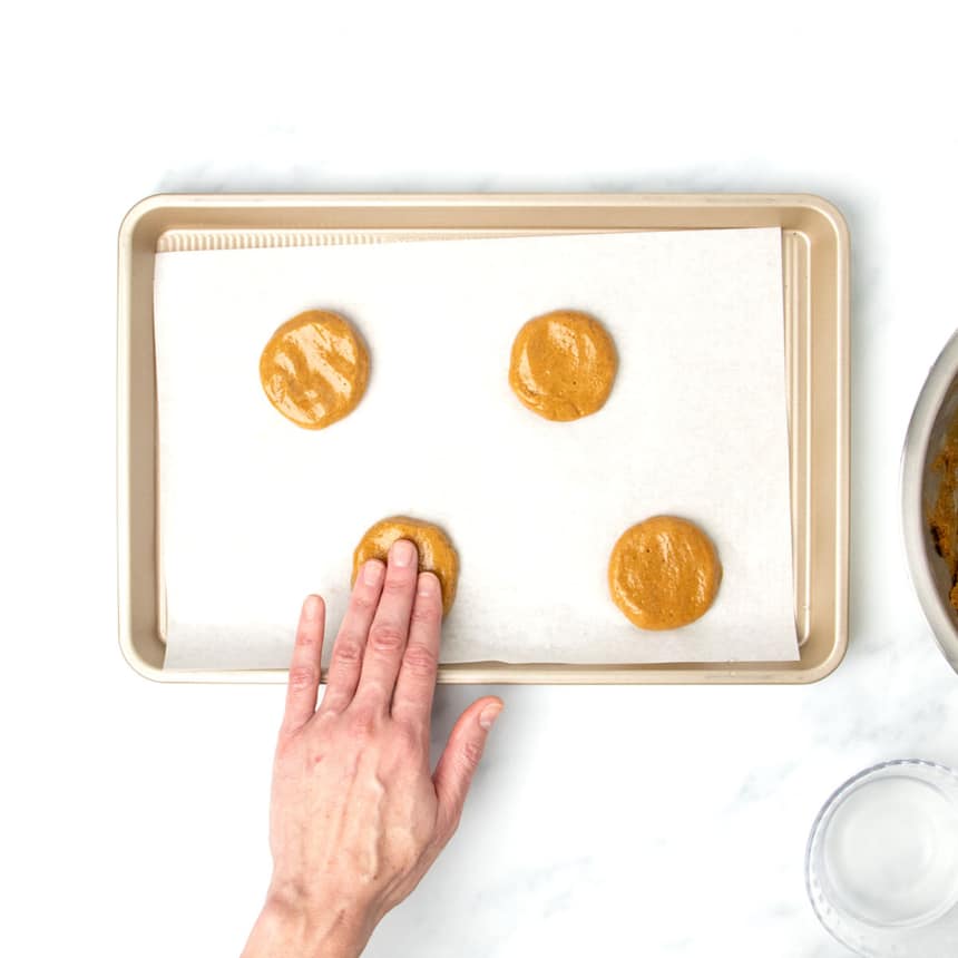 wet fingers pressing down on light brown round of cookie dough into a disk on white paper on gold baking tray