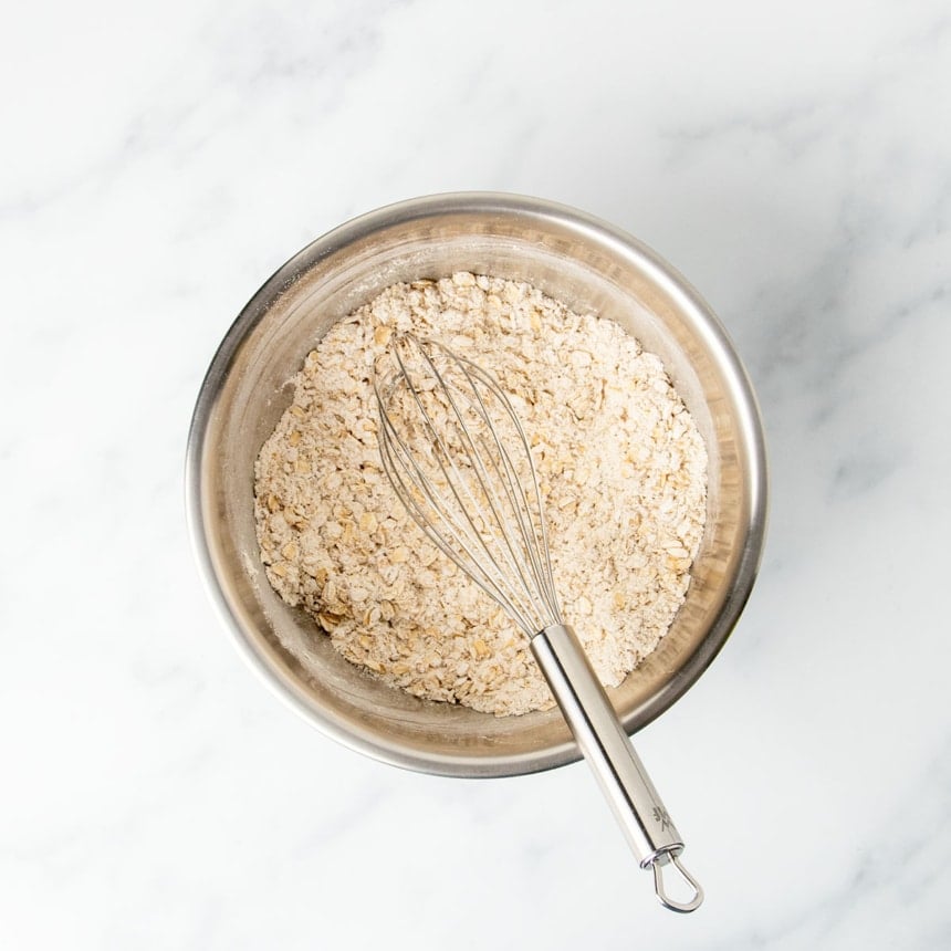 Medium size round metal mixing bowl with gluten free quick oats, oat flour and a large wire whisk