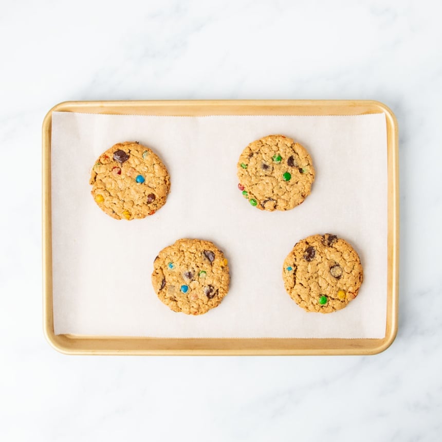 4 round light brown baked gluten free monster cookies with colored mini M&Ms candies and chocolate chips on same baking sheet