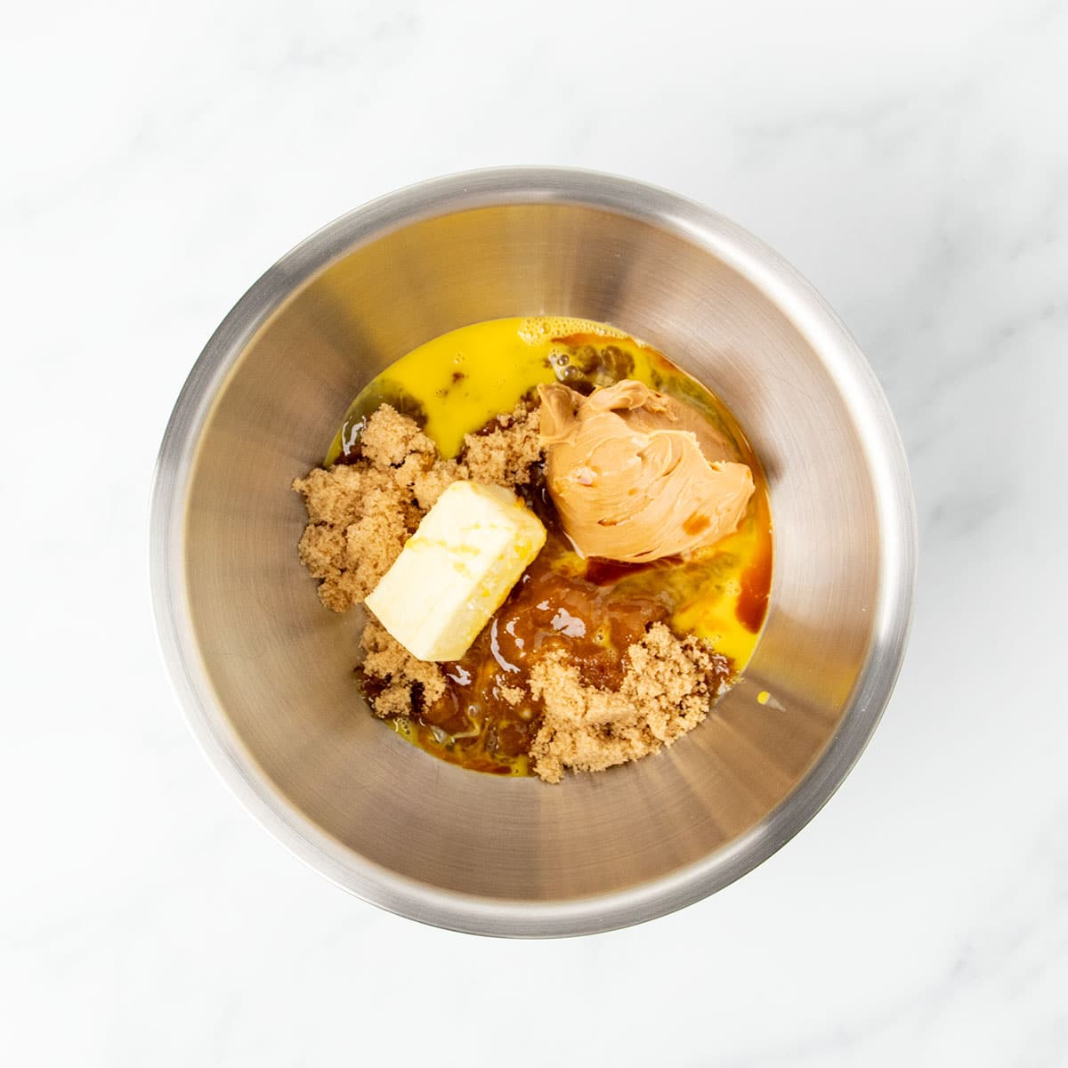 Large round metal mixing bowl with softened butter, gluten free peanut butter, brown sugar, beaten eggs, and vanilla