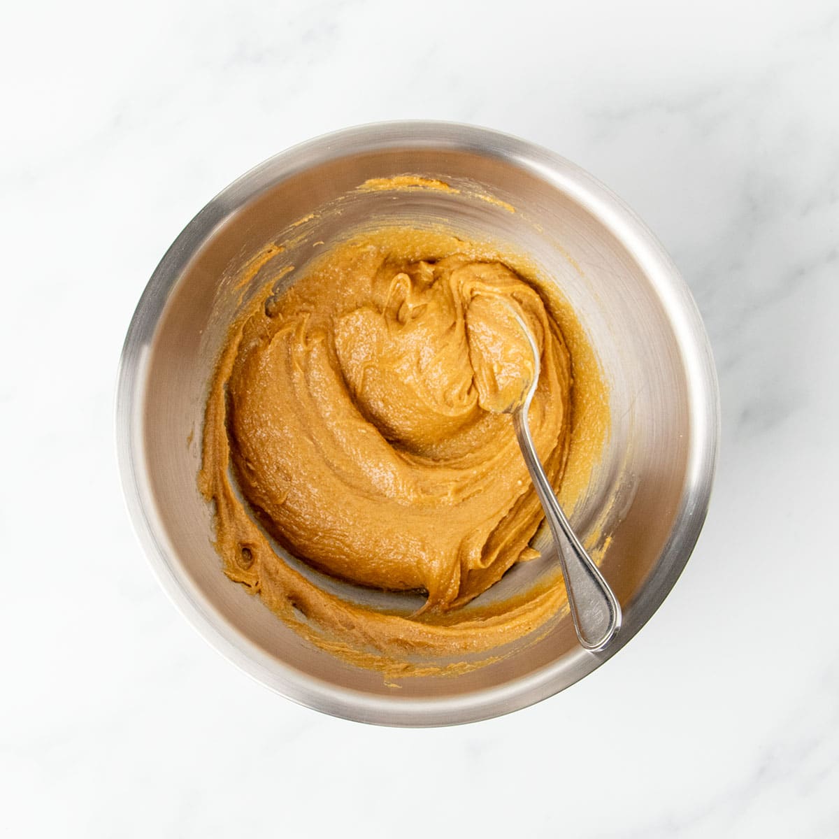 Same metal mixing bowl with large metal mixing spoon and mixed wet ingredients into a soft peanut butter colored mixture