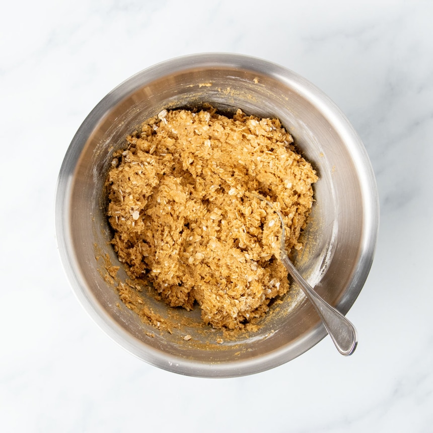 Same metal mixing bowl with oats and oat flour mixed into peanut butter mixture to form a thick light brown pasty cookie dough with a metal mixing spoon