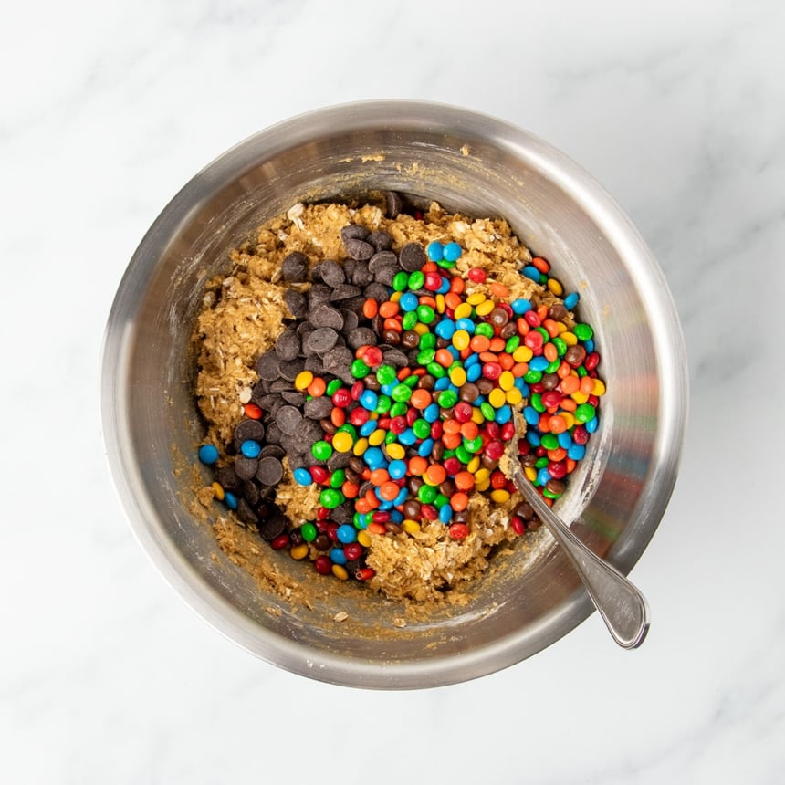 Same mixture in same bowl with mini M&Ms colored candies and chocolate chips sitting on top of dough