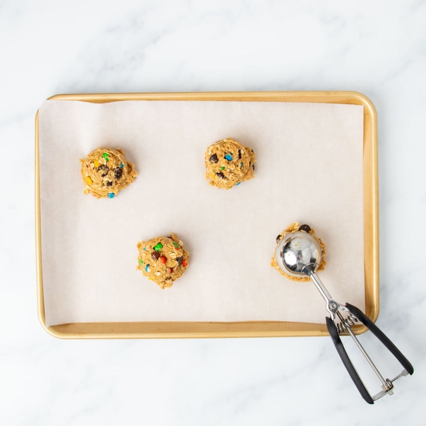 4 mounds of gluten free monster cookie dough with medium spring-loaded ice cream scoop sitting on one mound, all on a white paper-lined gold colored rimmed baking sheet