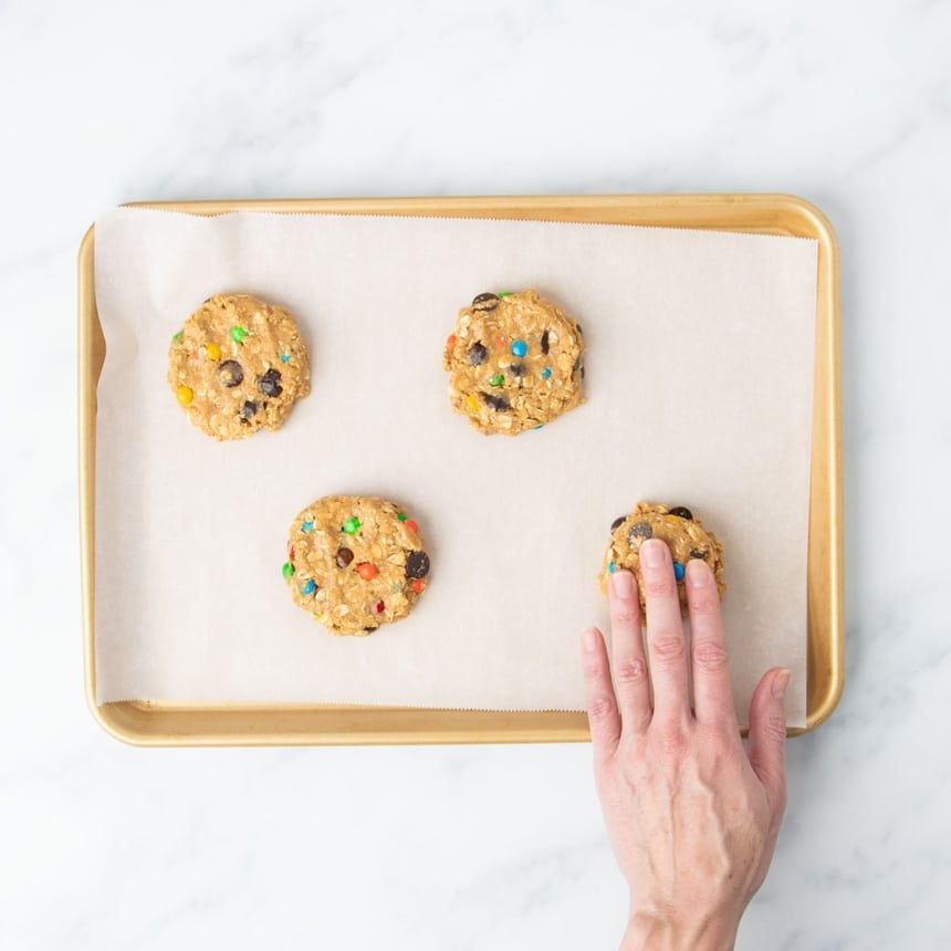 4 flat disks of monster cookie dough with fingers of one hand pressing down one piece of dough on same lined baking sheet