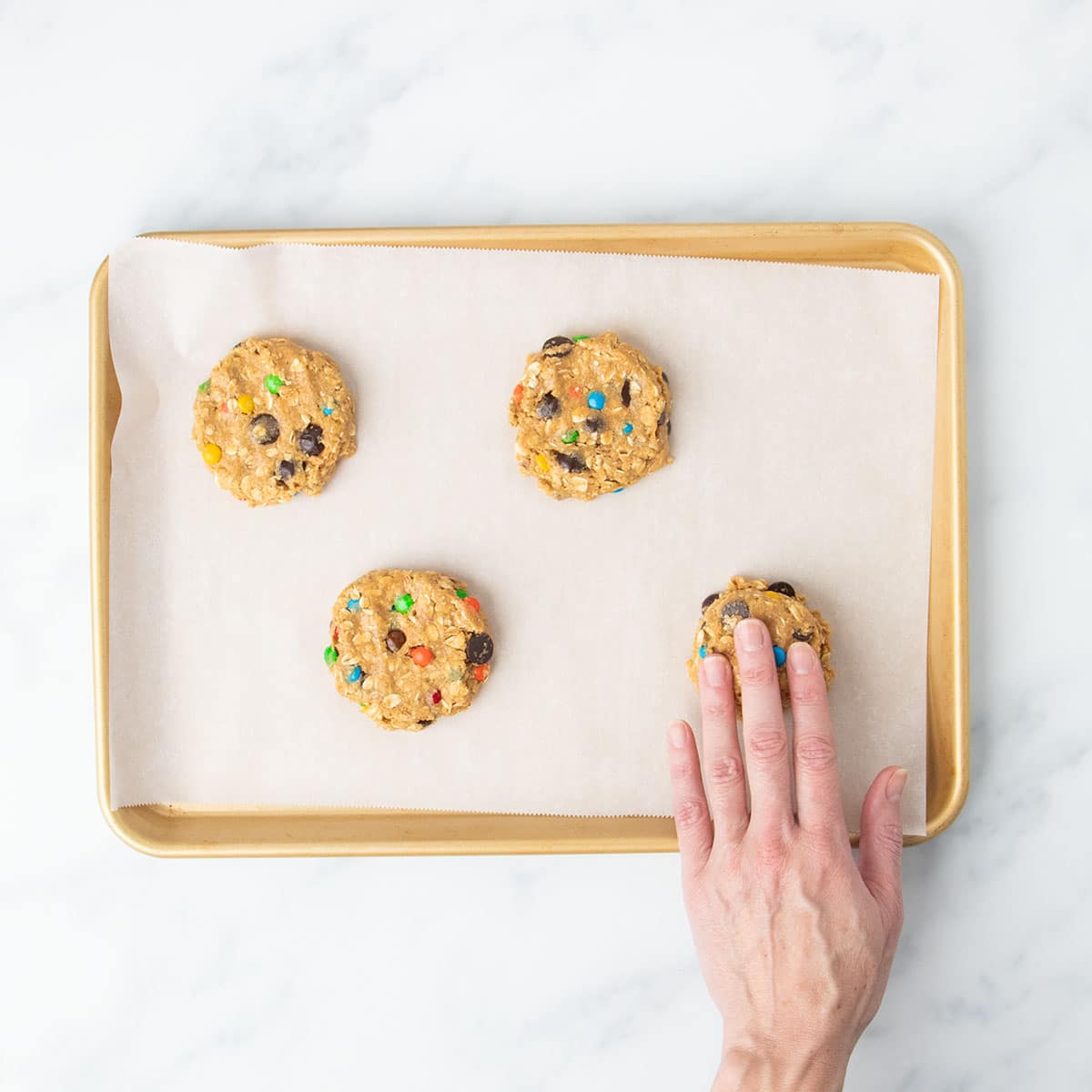 4 flat disks of monster cookie dough with fingers of one hand pressing down one piece of dough on same lined baking sheet