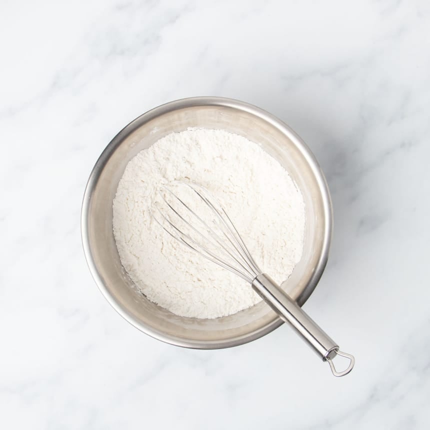 Round metal mixing bowl with white flour and wire whisk