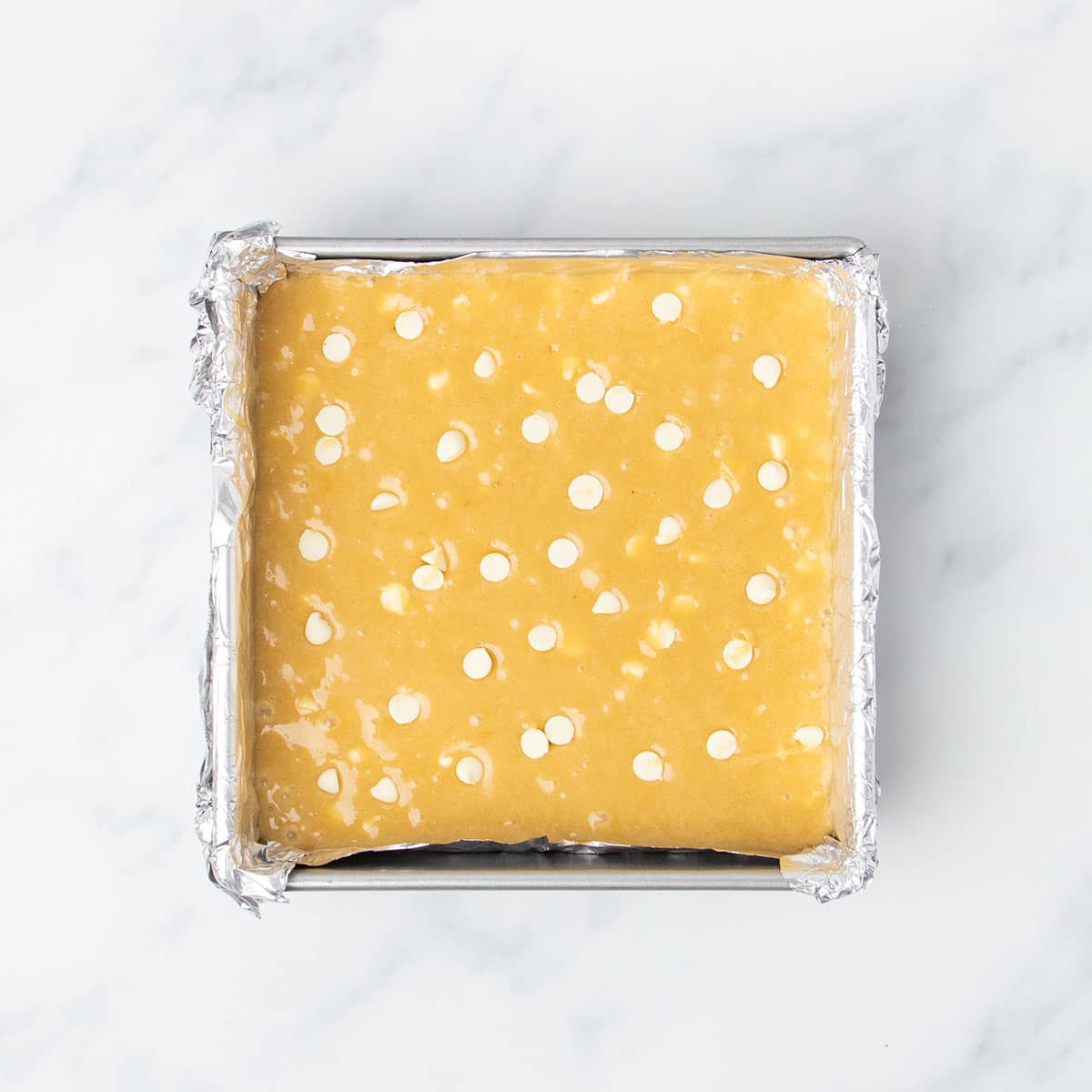 light brown raw blondie batter with white chocolate chips on top in foil-lined square metal baking pan