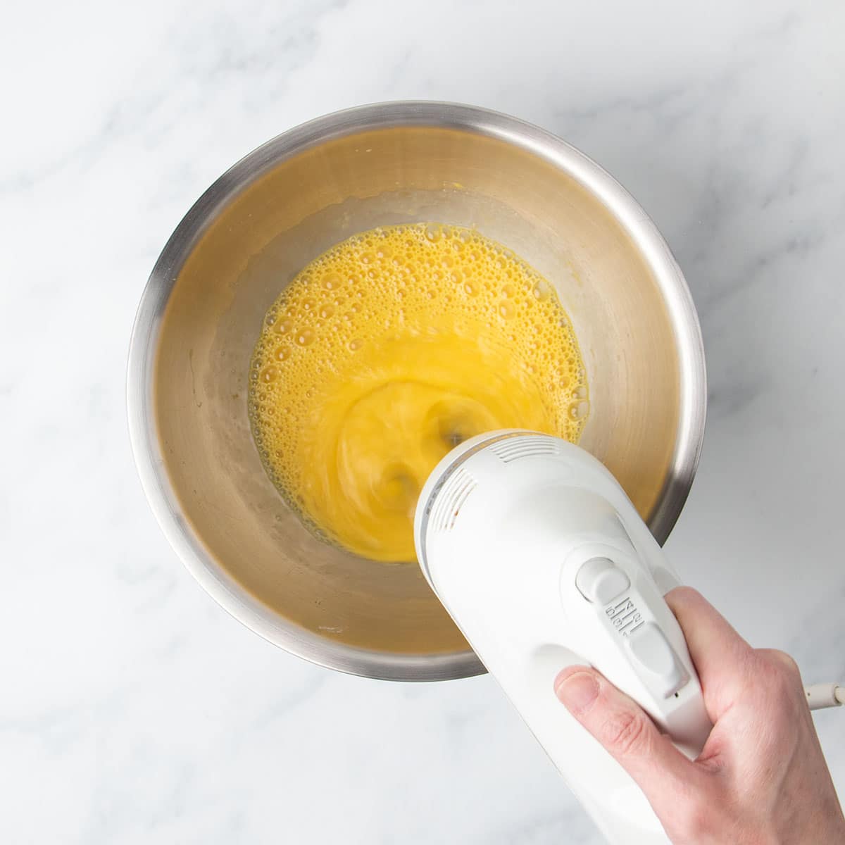 Hand holding white handheld mixer beating yellow egg mixture in large round metal mixing bowl