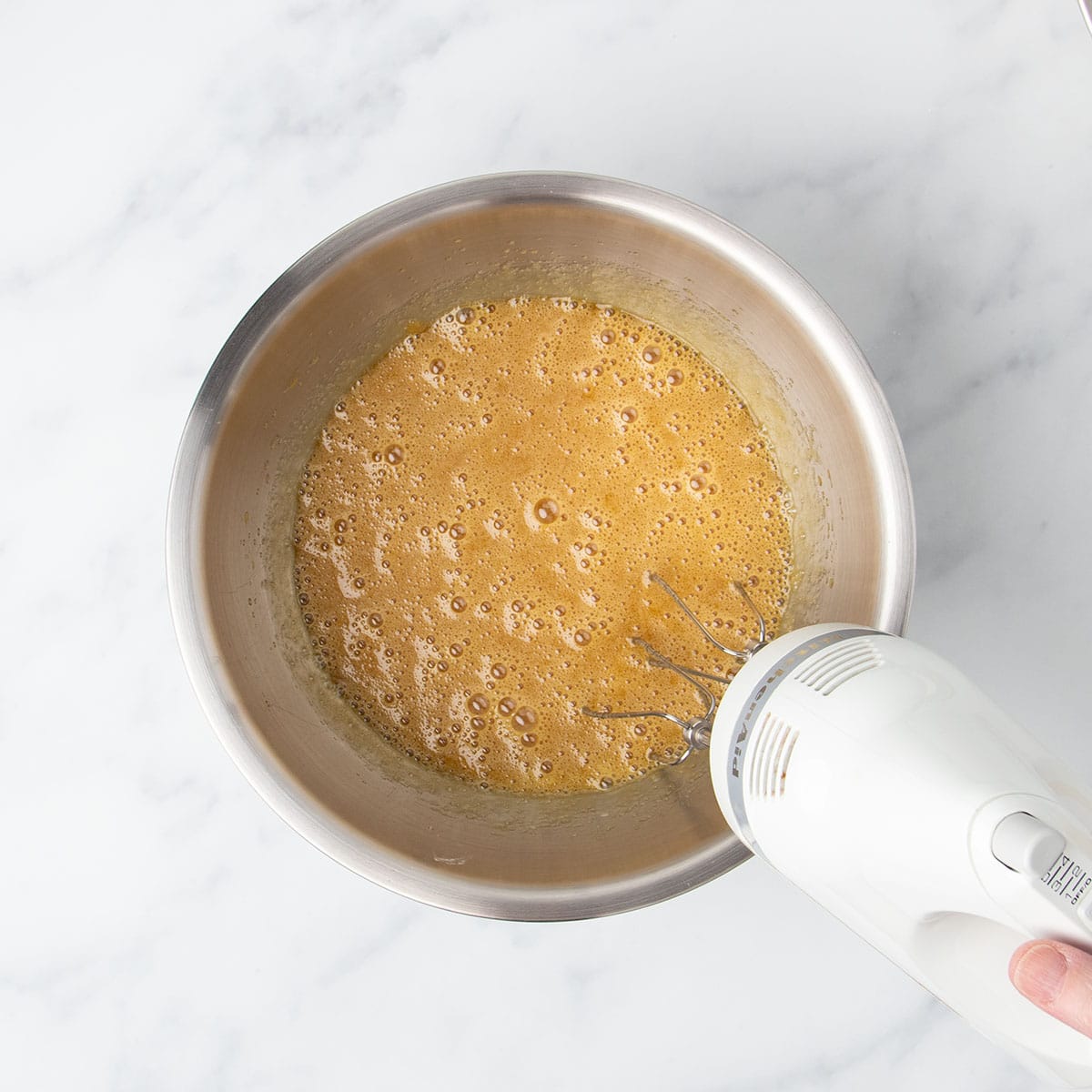 foamy light brown liquid mixture in large mixing bowl with hand holding white handheld mixer