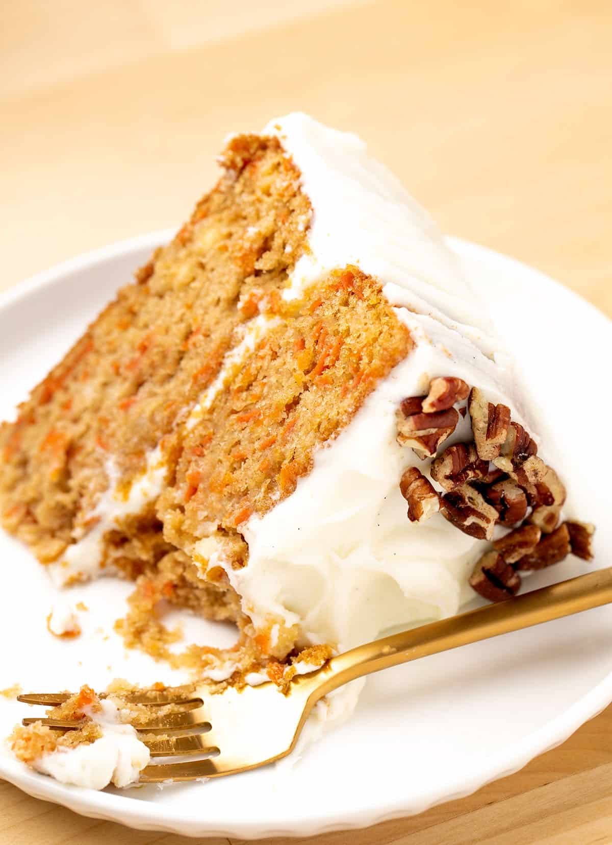 Slice of gluten free carrot cake with white frosting in between layers and chopped pecans on top on white plate with gold fork