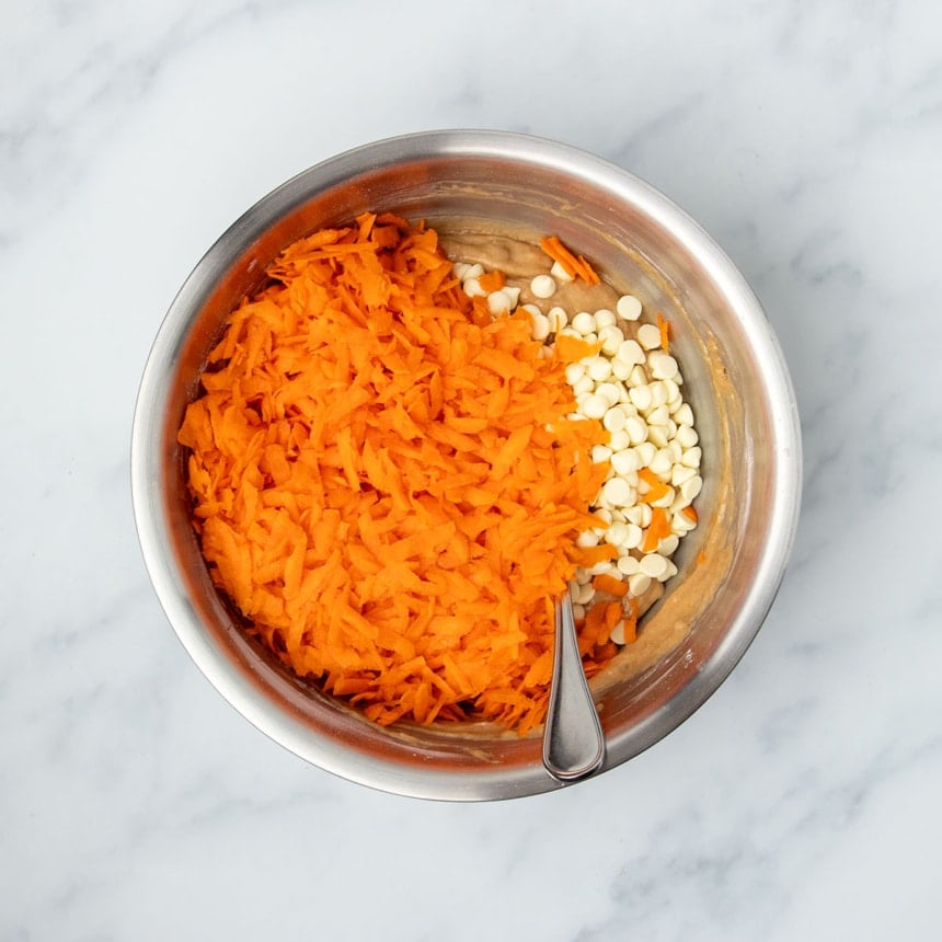 Same mixing bowl with light brown batter underneath and shredded orange carrots and white chocolate chips on top with metal mixing spoon