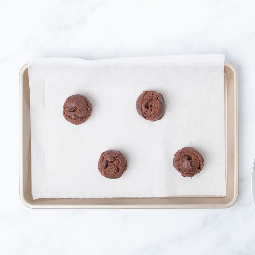 4 large round scoops of dark brown cookie dough spaced evenly apart on white paper on gold tray