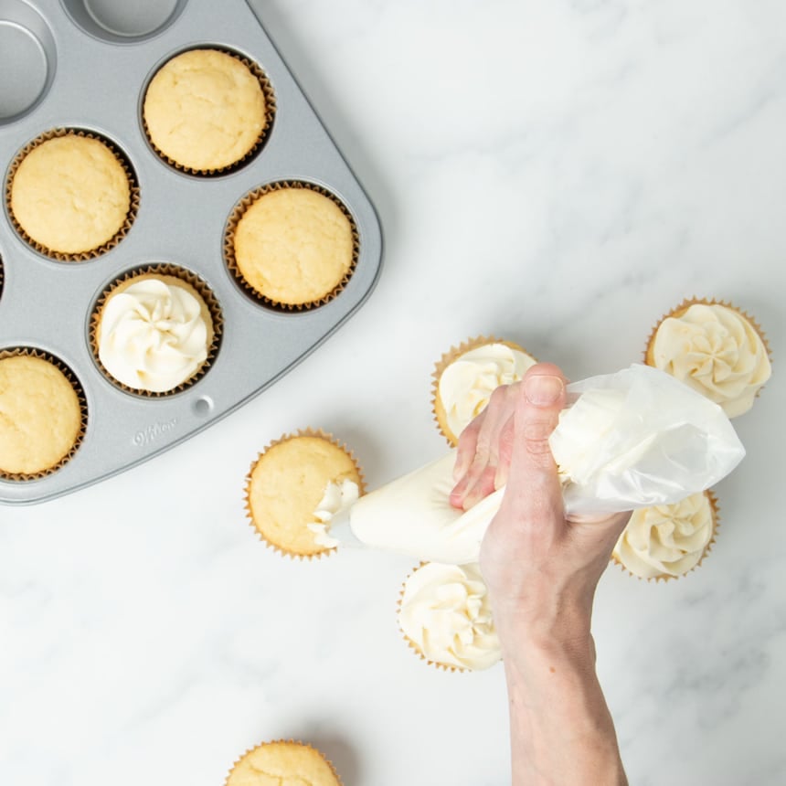 hand holding clear pastry bag with white frosting squeezing from a metal piping tip onto a yellow vanilla cupcake