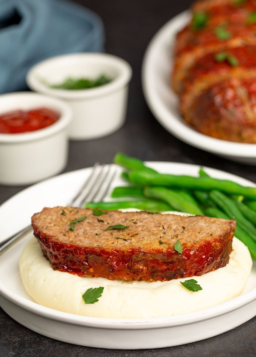 round white plate with slice of light brown meatloaf with red glaze on mound of light yellow mashed potatoes and green beans