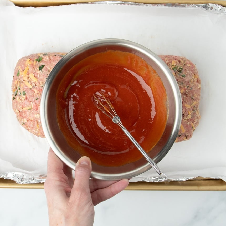 hand holding small round metal mixing bowl with red ketchup mixture and very small wire whisk