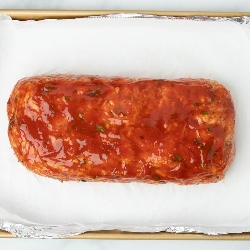 red ketchup glaze on top of shaped raw meatloaf on baking pan