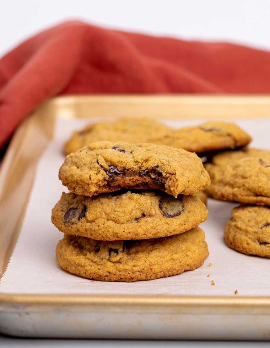 Brown-orange gluten free pumpkin chocolate chip cookies on a baking tray lined with white paper with a stack of 3 cookies in the front, the top one with a bite taken