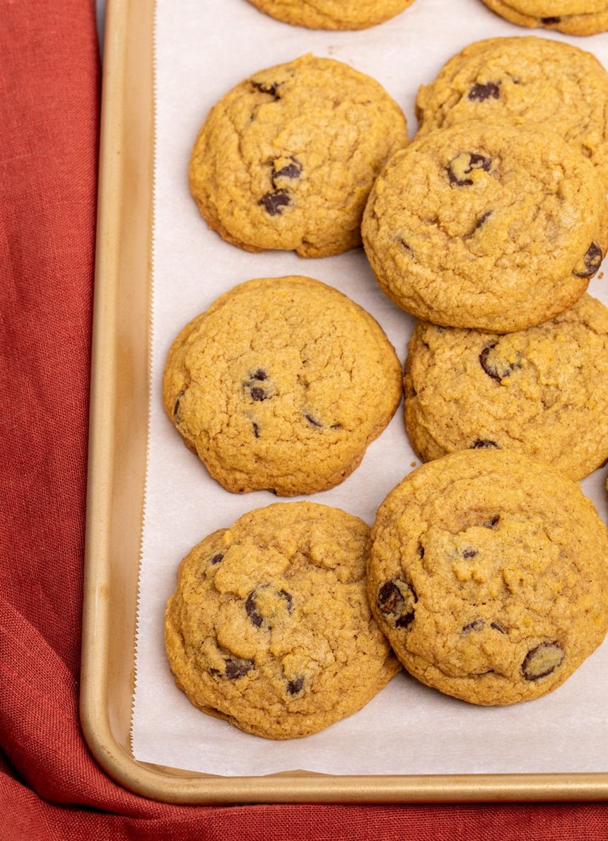 overhead image of 7 round cookies with chocolate chips and a light orange color on a gold baking sheet lined in white paper with a red cloth