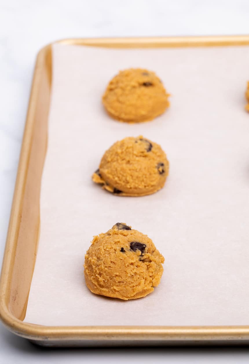 3 mounds of light orange balls of raw cookie dough with chocolate chips spaced evenly apart on white paper on baking tray