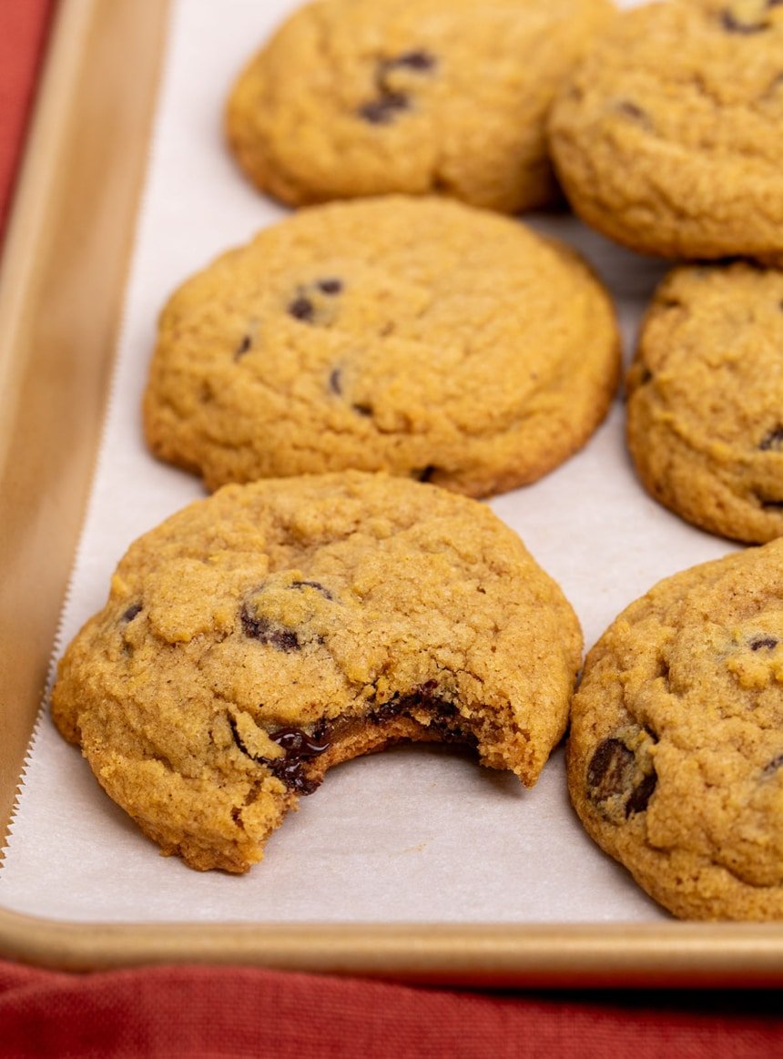 light brown-orange cookies with chocolate chips on baking tray with bite taken out of front cookie showing melted chocolate chips in center