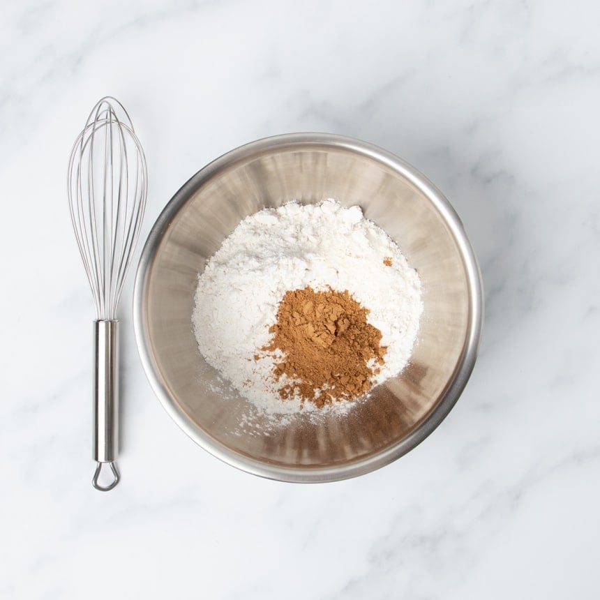 Small round metal mixing bowl with mostly white powder and some brown powder with wire whisk next to bowl